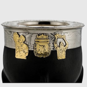 Puede incluir: Una calabaza de yerba mate negra con borde plateado y banda decorativa. La banda presenta figuras en relieve doradas y un emblema central. La calabaza está diseñada para el consumo tradicional de té sudamericano.