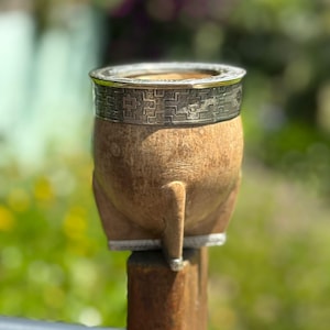 May include: A traditional yerba mate gourd, crafted from natural wood and adorned with a silver-toned metal band. The band features a detailed geometric design. The gourd has a distinctive shape and is supported by a small foot.