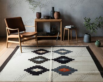 May include: A white and beige area rug with a geometric pattern in black, gray, and orange. The rug has a fringe border and is laid on a wooden floor. A brown leather armchair, a wooden table with a vase and plants, and a wooden stool are in the background.