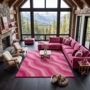 May include: A rectangular area rug featuring a wavy, multi-tonal pink pattern. The rug is positioned on a dark wooden floor in a living room with a large pink sectional sofa, two wooden armchairs with white cushions, and a fluffy cat sitting nearby.