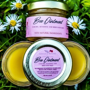 May include: Two small jars of Bee Ointment, a natural skin care product. The jars are gold with pink labels that read "Bee Ointment" and "FOR DRY, SENSITIVE AND IRRITATED SKIN". The jars are sitting on a bed of green grass and white daisies.