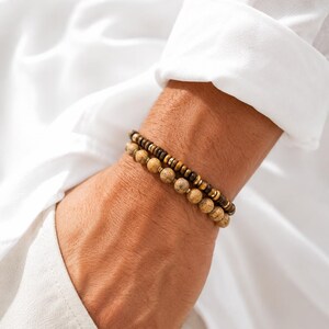 Pode incluir: Um conjunto de duas pulseiras de contas no pulso. Uma pulseira apresenta pequenas contas castanhas escuras em forma de disco com detalhes metálicos. A segunda pulseira consiste em contas de pedra maiores, redondas e com manchas castanhas.