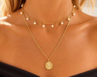 May include: A gold-toned layered necklace featuring two chains. The shorter chain is adorned with small white spherical pearls spaced evenly along the length. The longer chain features a circular sunburst pendant with a central sparkling stone.