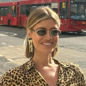 May include: A woman wearing a leopard print shirt and sunglasses with gold rectangular earrings. She is standing in front of a red double-decker bus.