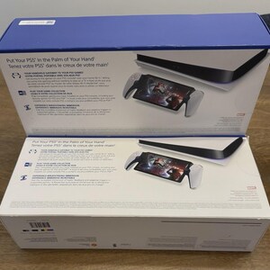 May include: Two white and blue PS5 boxes are open, revealing a console and a handheld device with a screen displaying a game. The boxes have text in English and French. The handheld device has white handles and a black screen.