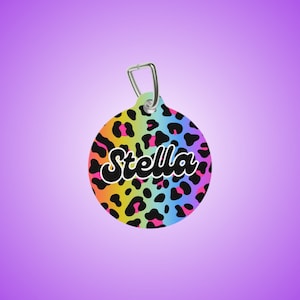 May include: A round, rainbow-colored leopard print dog tag with a silver ring and the name "Stella" in black cursive lettering.