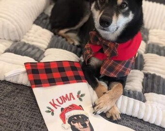Op de afbeelding: Een witte kerstsok met een rood-zwarte geruite manchet. De sok is geborduurd met de naam "KUMA" en een foto van een zwart-bruine hond met een kerstmanmuts.