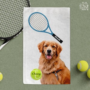 May include: A white towel featuring a golden retriever with a tennis racket and tennis ball. The racket is blue with the name "Sarah" on the handle. The tennis ball has the name "Chewy". Two yellow tennis balls are also visible.