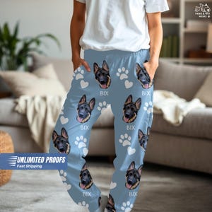 May include: Light blue pajama pants featuring a repeating pattern of German Shepherd dog faces, white paw prints, and hearts. The word "BIX" is printed below each dog face. The pants have pockets and are made of a soft material.