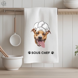 May include: White waffle-weave kitchen towel with a smiling dog wearing a chef's hat, the name "Zeus" on it. The text "SOUS CHEF" and paw prints are printed below the dog image. A wooden spoon, ladle, and bowl are in the background.