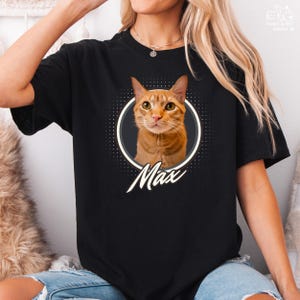 以下が含まれることがあります： オレンジ色のトラ猫、マックスの肖像画が描かれた黒いTシャツ。猫の画像は、暗い灰色の境界線を持つ白い点線円の中にあります。「Max」という名前が猫の画像の下に筆記体で書かれています。