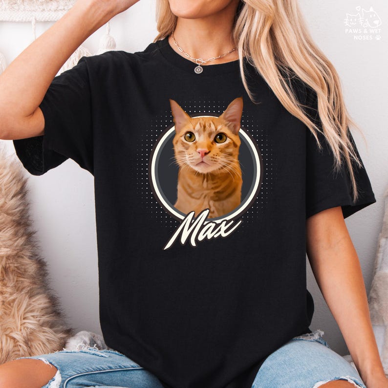 以下が含まれることがあります： オレンジ色のトラ猫、マックスの肖像画が描かれた黒いTシャツ。猫の画像は、暗い灰色の境界線を持つ白い点線円の中にあります。「Max」という名前が猫の画像の下に筆記体で書かれています。