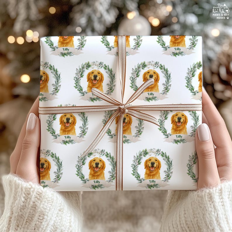 Op de afbeelding: Wit cadeaupapier met een herhalend patroon van een Golden Retriever-gezicht in een groene krans, met de naam "Lilly" eronder. Het cadeau is vastgebonden met een bruin lint.
