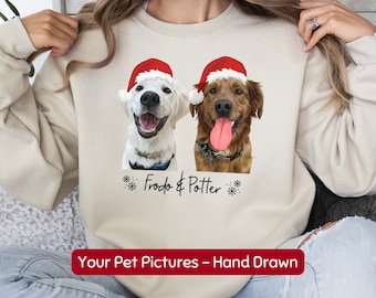 May include: A beige sweatshirt with a hand-drawn illustration of two dogs wearing Santa hats. The dogs are a white Golden Retriever and a golden retriever. The text "Frodo & Potter" is printed below the dogs.