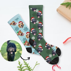 May include: Two pairs of Christmas-themed socks. One pair is light blue with dog faces wearing Santa hats and snowflakes. The other pair is green with black dog faces wearing Santa hats and snowflakes. The socks are a fun and festive holiday gift.