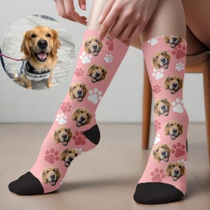 May include: Pink socks featuring a repeating pattern of a golden retriever's face and paw prints. The socks have black heels and toes. A circular image of a golden retriever is in the background.