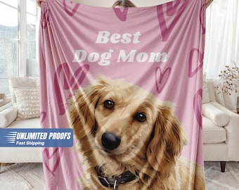 May include: A pink blanket featuring a golden retriever with the text "Best Dog Mom" in white. The blanket has heart-shaped designs and a close-up of the dog's face. The blanket is held up by a person.