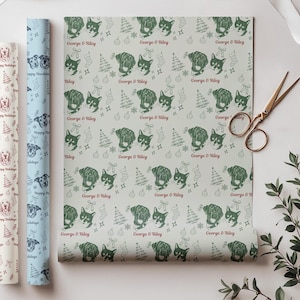 May include: A roll of light gray wrapping paper featuring a repeating pattern of dark green dog portraits, Christmas trees, stockings, and ornaments, with the red text George & Riley printed throughout. A pair of metal scissors lies nearby.