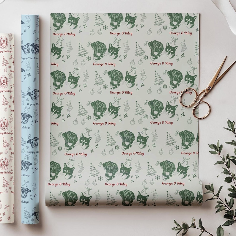 May include: A roll of light gray wrapping paper featuring a repeating pattern of dark green dog portraits, Christmas trees, stockings, and ornaments, with the red text George & Riley printed throughout. A pair of metal scissors lies nearby.