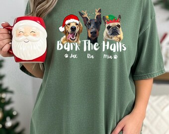 Puede incluir: Una camiseta verde con la frase "Bark The Halls" y tres perros con accesorios navideños. Un golden retriever lleva un gorro de Papá Noel, un Doberman tiene cuernos y un bulldog francés lleva un gorro de elfo. Una taza roja de Papá Noel se sostiene en la mano izquierda.