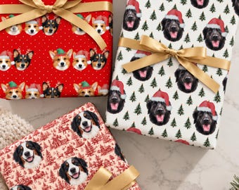 Op de afbeelding: Drie rechthoekige cadeaus verpakt in bedrukt papier met gouden linten. Eén rood papier toont Corgi-gezichten met kerstmutsen, één wit papier toont zwarte honden met kerstmutsen en dennenbomen, en één crèmekleurig papier toont Berner Sennenhonden.