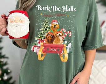 Puede incluir: Camiseta verde con el texto "Bark The Halls" y "Santa's Helpers". La camiseta presenta una ilustración de acuarela de tres perros en un trineo rojo con un árbol de Navidad, bastones de caramelo y regalos envueltos. Una taza roja de Papá Noel se sostiene en la mano izquierda.