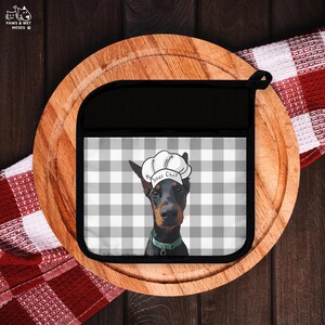 May include: Black and gray pot holder with a Doberman Pinscher wearing a chef's hat that says "Sous Chef". The background is a gray and white checkered pattern. The pot holder is on a wooden cutting board.