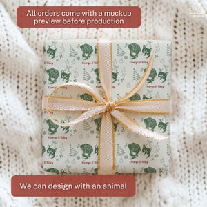 May include: A square gift box wrapped in light-colored paper featuring a repeating dark green pattern of dog and cat faces, Christmas trees, stockings, and ornaments, with the text George & Riley, tied with a shimmering gold ribbon.