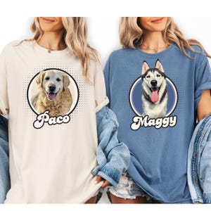 以下が含まれることがあります： クリーム色と青色の2枚のTシャツ。それぞれ円の中に犬の肖像画が描かれています。クリーム色のシャツには「Paco」という名前のゴールデンレトリバーが描かれています。青色のシャツには「Maggy」という名前のハスキーが描かれています。
