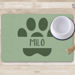 May include: A sage green pet feeding mat with a dark green paw print and the name "MILO". A white and navy blue pet bowl sits on the mat, with a bone-shaped dog treat nearby. The mat is on a wooden surface.