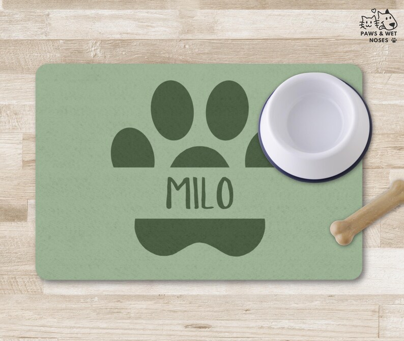 May include: A sage green pet feeding mat with a dark green paw print and the name "MILO". A white and navy blue pet bowl sits on the mat, with a bone-shaped dog treat nearby. The mat is on a wooden surface.