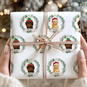 Op de afbeelding: Wit cadeaupapier met herhalende patronen van een bruine hond en een oranje kat, elk met een kerstmuts en omringd door een krans. De hond is gelabeld met "Maggy" en de kat met "Reggie". Een gouden lint is om het cadeau gebonden.