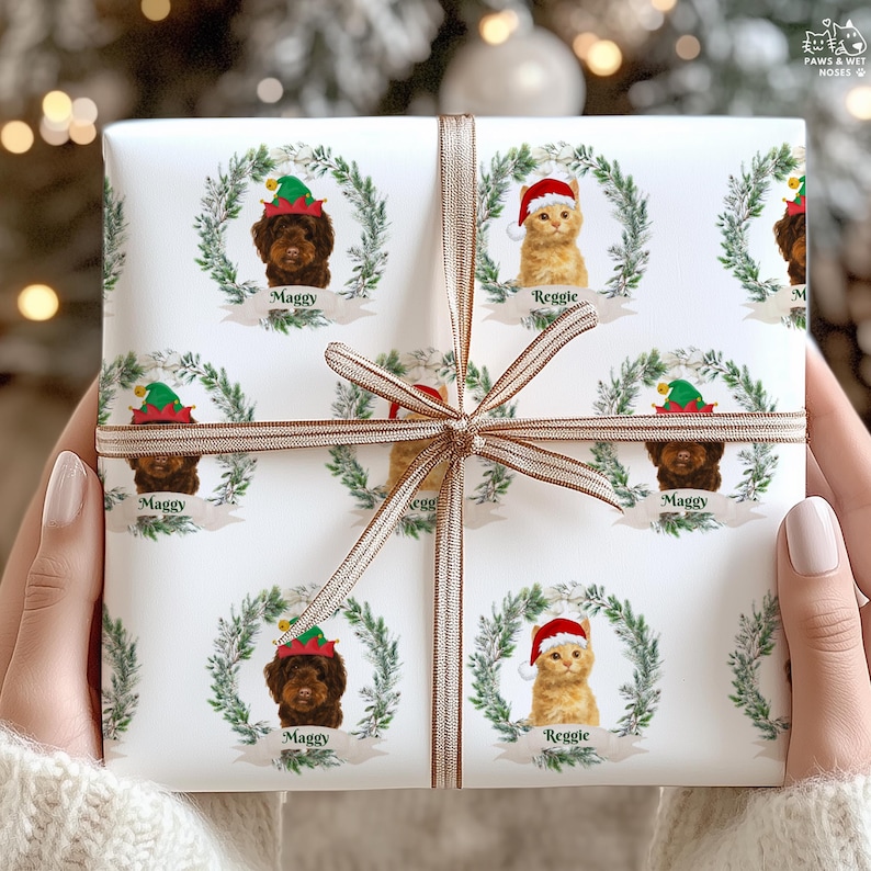 Op de afbeelding: Wit cadeaupapier met herhalende patronen van een bruine hond en een oranje kat, elk met een kerstmuts en omringd door een krans. De hond is gelabeld met "Maggy" en de kat met "Reggie". Een gouden lint is om het cadeau gebonden.