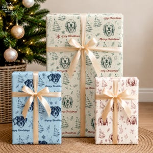 May include: Three rectangular gift boxes wrapped in patterned paper and tied with champagne-colored satin ribbons. The large center box is cream with green dog illustrations and Merry Christmas text. The blue and white boxes feature dog faces.