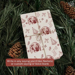 May include: A rectangular gift wrapped in cream-colored paper featuring a repeating dark red line drawing pattern of dachshund faces, Christmas trees, stockings, bows, and ornaments, tied with thin brown twine string across the center.