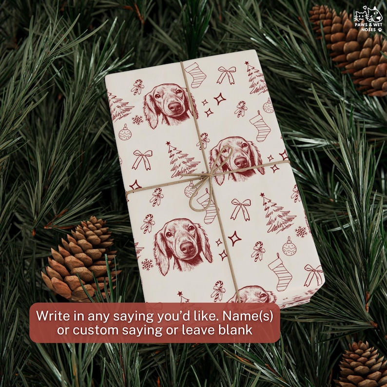 May include: A rectangular gift wrapped in cream-colored paper featuring a repeating dark red line drawing pattern of dachshund faces, Christmas trees, stockings, bows, and ornaments, tied with thin brown twine string across the center.