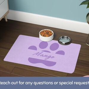May include: A lavender pet feeding mat with a paw print design and the name "Mango" in script. A white bowl filled with pet food and a silver bowl sit on the mat. The mat is on a dark wood floor.