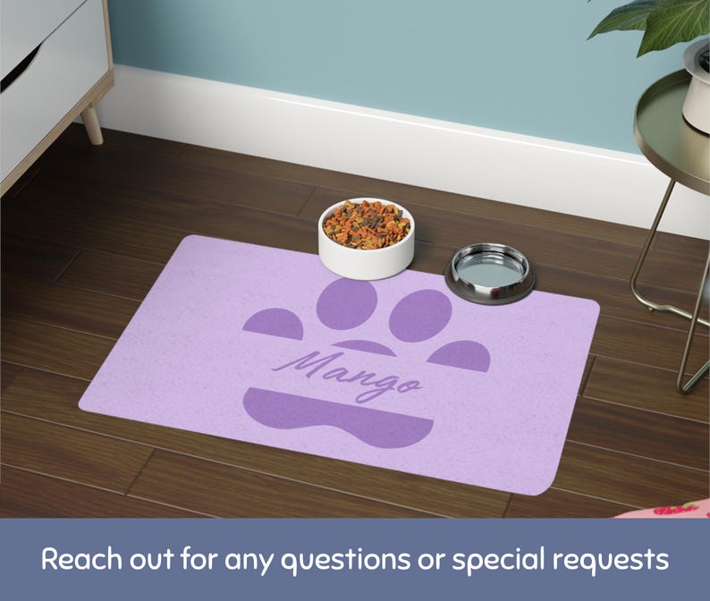 May include: A lavender pet feeding mat with a paw print design and the name "Mango" in script. A white bowl filled with pet food and a silver bowl sit on the mat. The mat is on a dark wood floor.