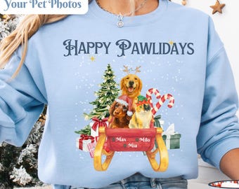 以下が含まれることがあります： 「Happy Pawlidays」と「Your Pet Photos」の文字が入った水色のスウェットシャツ。クリスマスツリー、プレゼント、キャンディケーンと一緒に犬がそりに乗っている水彩画イラストが特徴です。