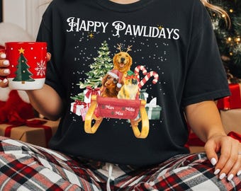 Puede incluir: Camiseta negra con el texto "Happy Pawlidays" en escritura blanca. La camiseta presenta un gráfico festivo de perros y un gato en un trineo con regalos, un árbol de Navidad y bastones de caramelo. Los nombres "Max", "Mia" y "Milo" están en el trineo.