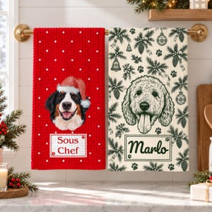 May include: Two waffle-weave kitchen towels hanging on a gold rail. The left towel is red with white polka dots, a Bernese Mountain Dog wearing a Santa hat, and a box labelled Sous Chef. The right towel is cream with a black pine and ornament pattern, a doodle dog, and a box labelled Marlo.
