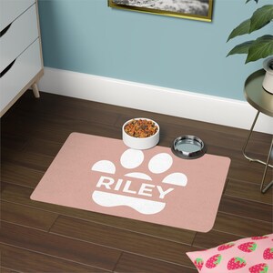 May include: A rectangular, blush-colored pet food mat with a white paw print design and the name "RILEY" in white block letters. A white ceramic bowl filled with pet food and a stainless steel bowl sit on the mat.