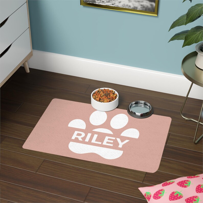 May include: A rectangular, blush-colored pet food mat with a white paw print design and the name "RILEY" in white block letters. A white ceramic bowl filled with pet food and a stainless steel bowl sit on the mat.