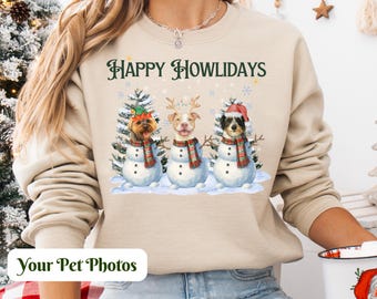 以下が含まれることがあります： ベージュ色のスウェットシャツ。緑色の文字で「HAPPY HOWLIDAYS」と書かれています。雪だるまに扮した3匹の犬が、スカーフと帽子を身につけています。「Your Pet Photos」の文字が画像の下部にあります。
