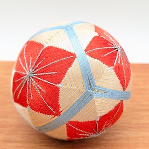 Op de afbeelding: Een decoratieve Temari-bal met een complex geometrisch ontwerp. De bal heeft rode, lichtblauwe en crèmekleurige borduursels. Het ontwerp omvat stervormige patronen en lineaire accenten, waardoor een visueel aantrekkelijk handgemaakt item ontstaat.