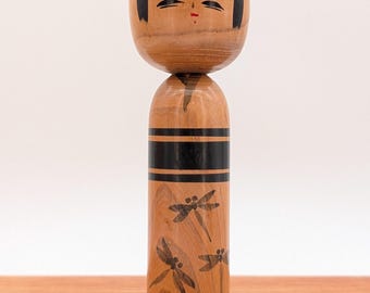 May include: A wooden kokeshi doll with a rounded head and cylindrical body. The head features painted black hair and facial details with a small red mouth. The body is decorated with horizontal black bands and black dragonfly illustrations.