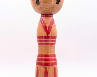May include: A wooden Kokeshi doll featuring a large, rounded head with painted black hair and facial features. The cylindrical body is decorated with red painted patterns, including horizontal bands and vertical stripes along the base.