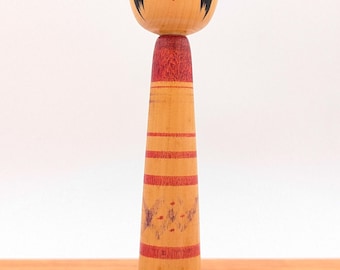 May include: A wooden Kokeshi doll featuring a rounded head with painted black hair, red floral accents, and a neutral facial expression. The cylindrical body is light brown with horizontal red stripes and a faded purple pattern in the center.