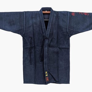 Op de afbeelding: Een donkerblauw, indigo-geverfd katoenen jasje met wijde mouwen en een V-hals. Het kledingstuk is voorzien van rode geborduurde Japanse karakters op de linkerschouder en gele Japanse tekst met een klein rood vierkant embleem op het voorpand.