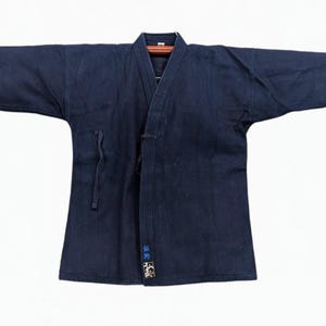 Op de afbeelding: Een donker marineblauw indigo-geverfd kendo-jasje met wijde mouwen, een V-hals met verstevigde stiksels en een overslagsluiting met stoffen linten. De onderste voorrand is voorzien van kleine, blauw geborduurde Japanse Kanji-tekens.
